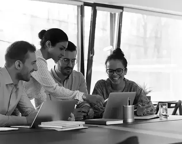 A diverse group of individuals sitting around a table with laptops, engaged in a meeting or collaboration session.